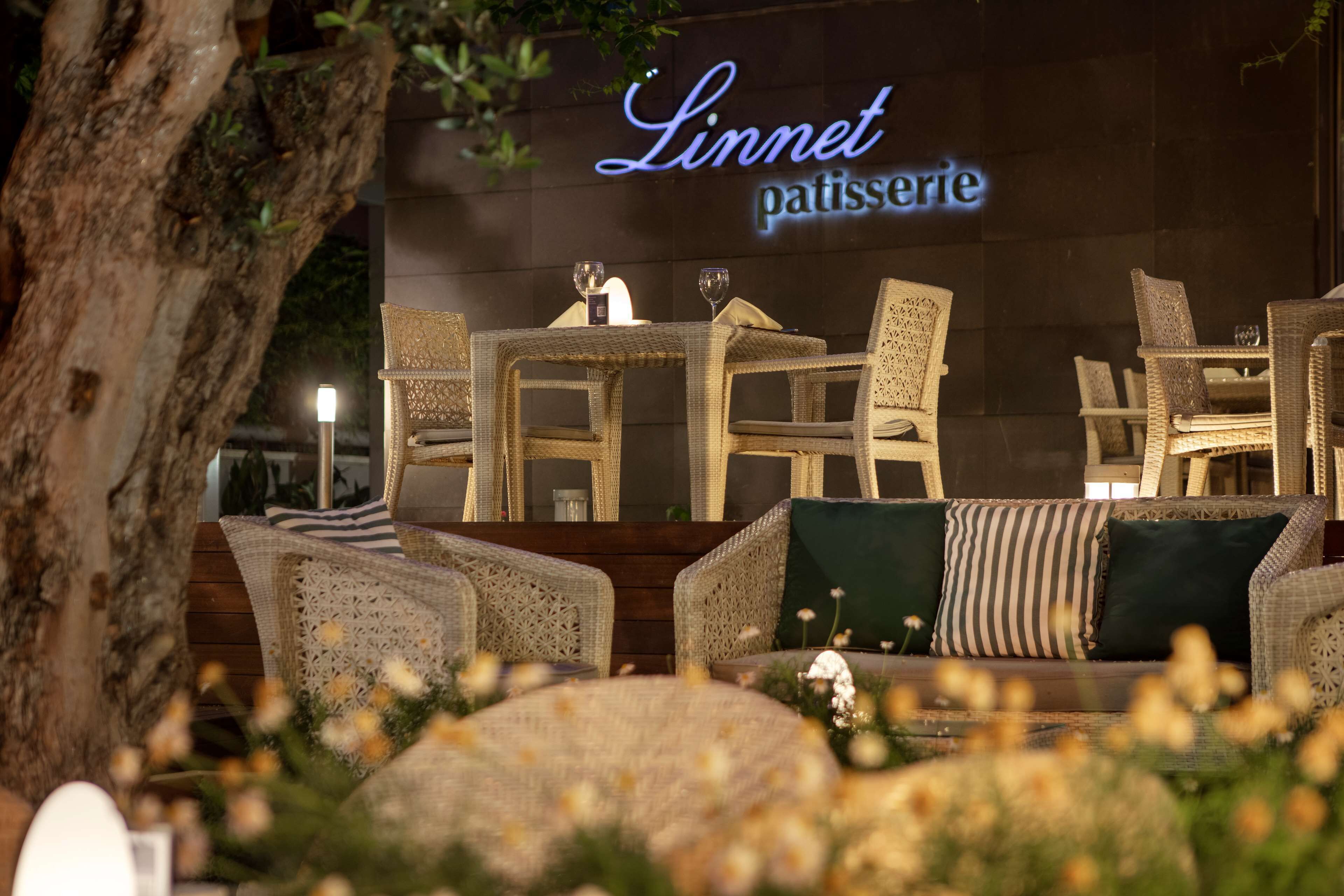 An outdoor seating area with wicker furniture, a sign reading "Linnet patisserie," and a tree in the background.