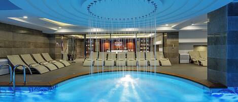 A modern indoor pool with a circular ceiling design, lounge chairs, and a reception area in the background.