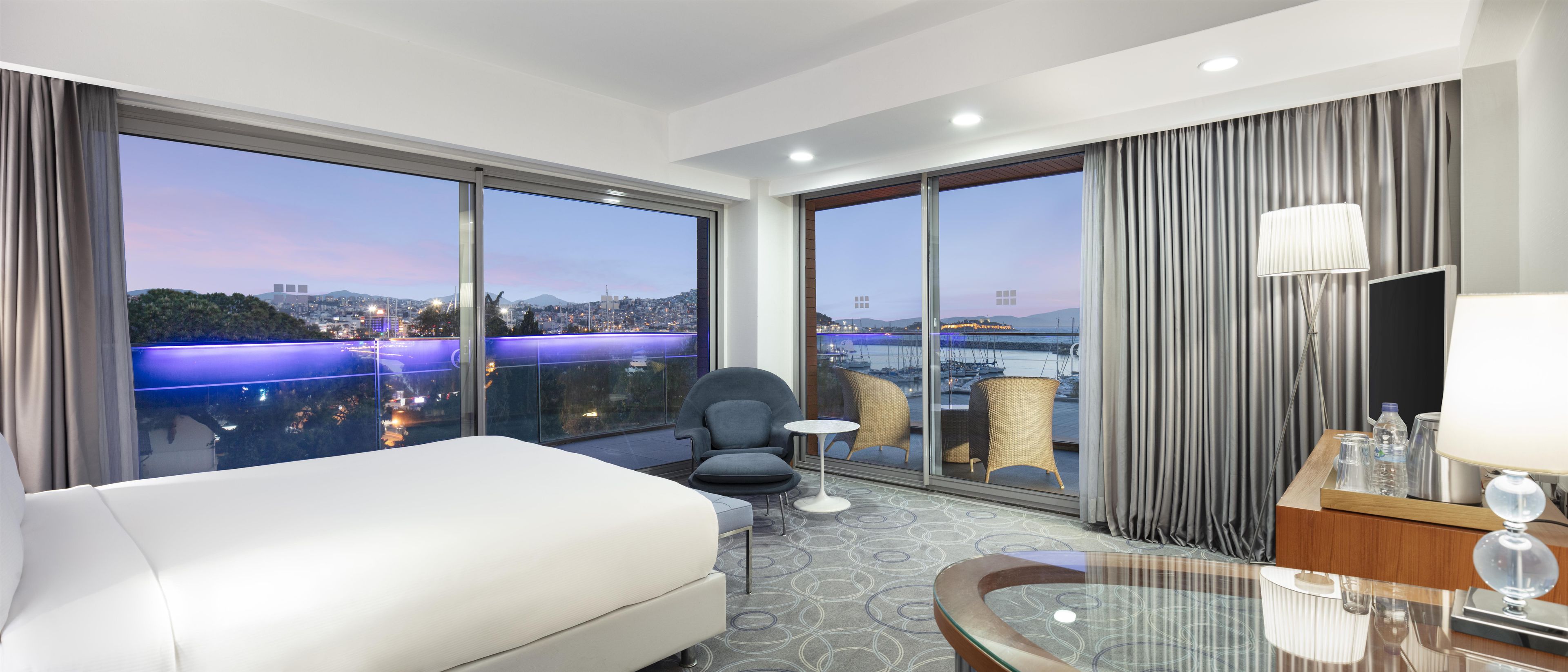 A hotel room with a large bed, a small table, a chair, a desk with a lamp, and a view of a cityscape and harbor.