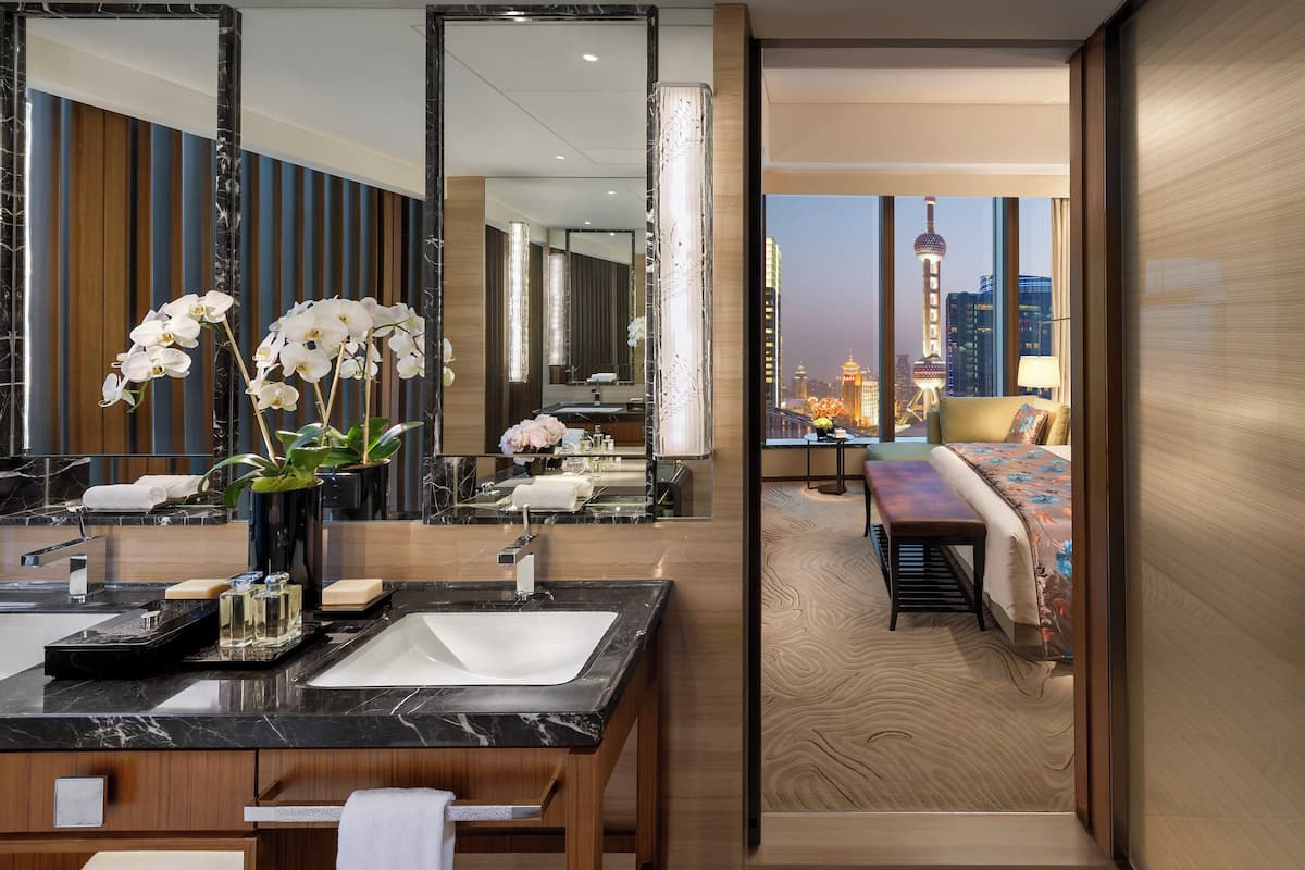 A modern hotel bathroom with a large mirror, marble countertop, and a view of the cityscape.