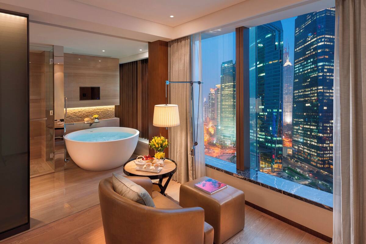 A modern hotel room with a large bathtub, a sofa, a small table with a vase of flowers, and a city view through large windows.