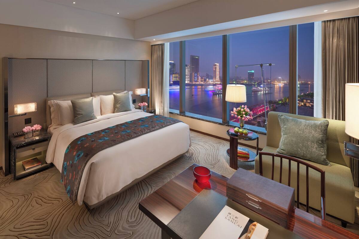 A modern hotel room with a large bed, a desk, and a view of a cityscape at night.