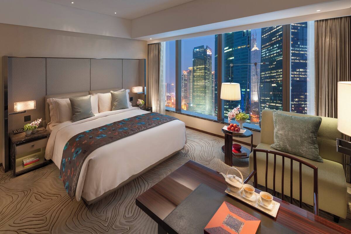 Een moderne hotelkamer met een groot bed, uitzicht op de stad, een kleine tafel met een dienblad thee en een comfortabele fauteuil.