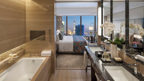 Una habitación de hotel moderna con una cama grande, un baño independiente con bañera, un tocador con espejo y vistas a la silueta urbana.