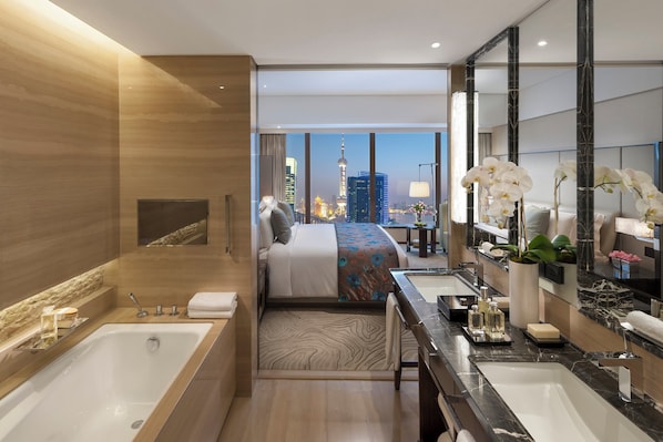 A modern hotel room with a large bed, a separate bathroom with a bathtub, a vanity with a mirror, and a view of a city skyline.