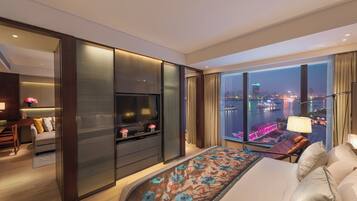 A modern hotel room with a large bed, a TV, a desk, and a view of the cityscape at night.