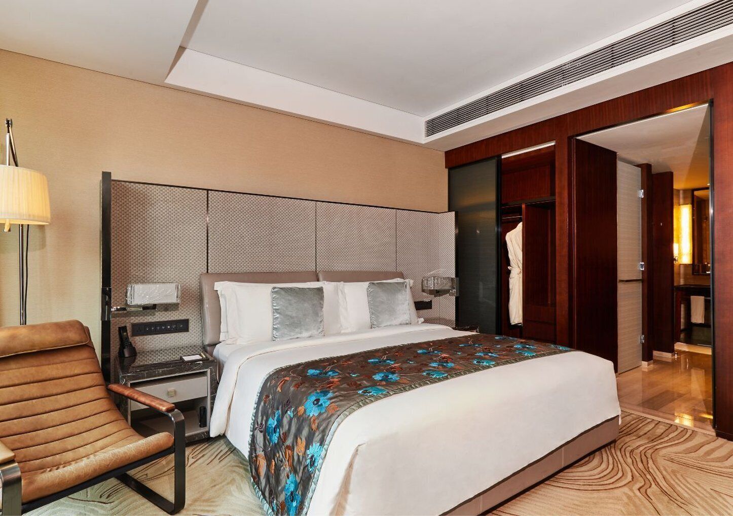 A modern hotel room with a large bed, a desk with a lamp, a leather chair, and a walk-in closet.