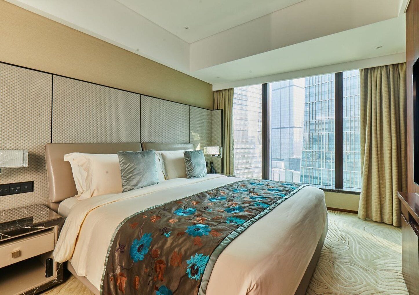 Habitación de hotel moderna con una cama grande, una mesita de noche, un televisor y vistas de la ciudad a través de amplios ventanales.