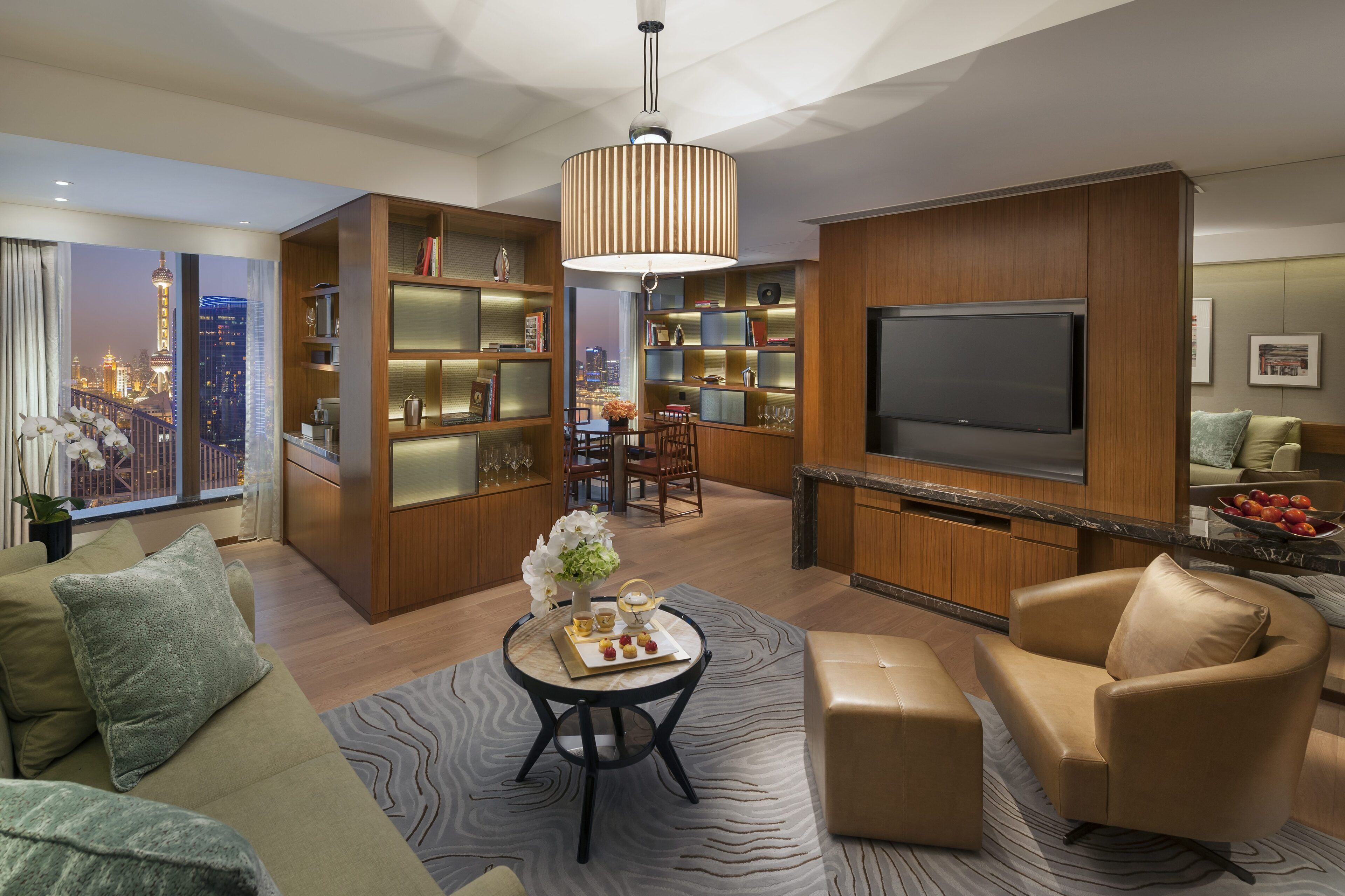 A modern living room with a large TV, wooden shelving, and a dining area with a city view.