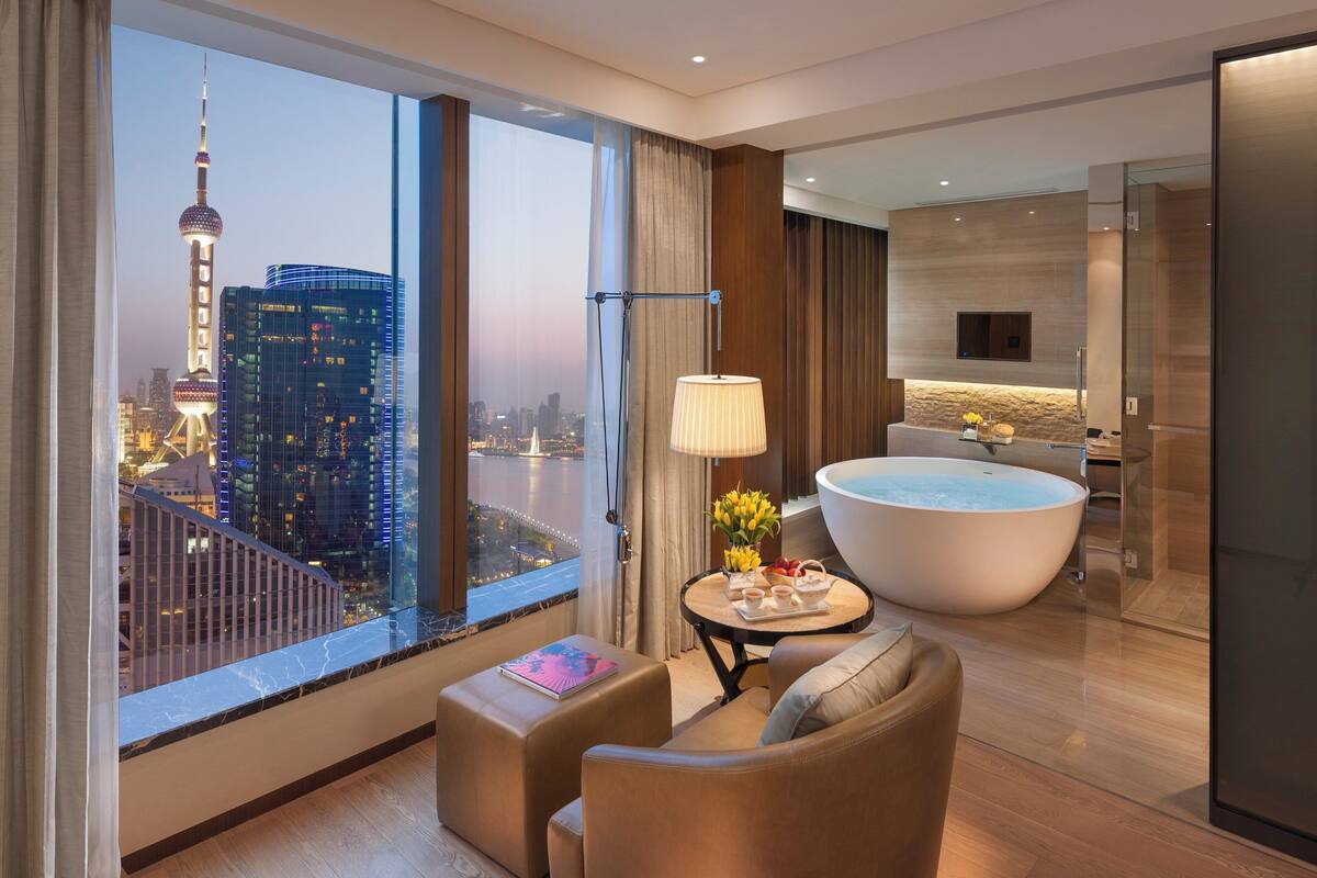 Een moderne hotelkamer met een groot raam met uitzicht op de stad, een zithoek met een tafel en stoelen, een badkuip en een kleine keukenhoek.