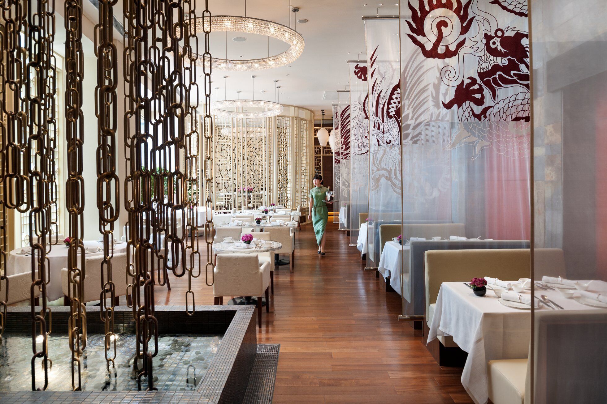 A restaurant with a modern interior, featuring a long dining area with tables and chairs, a decorative wall with intricate patterns, and a hanging light fixture.
