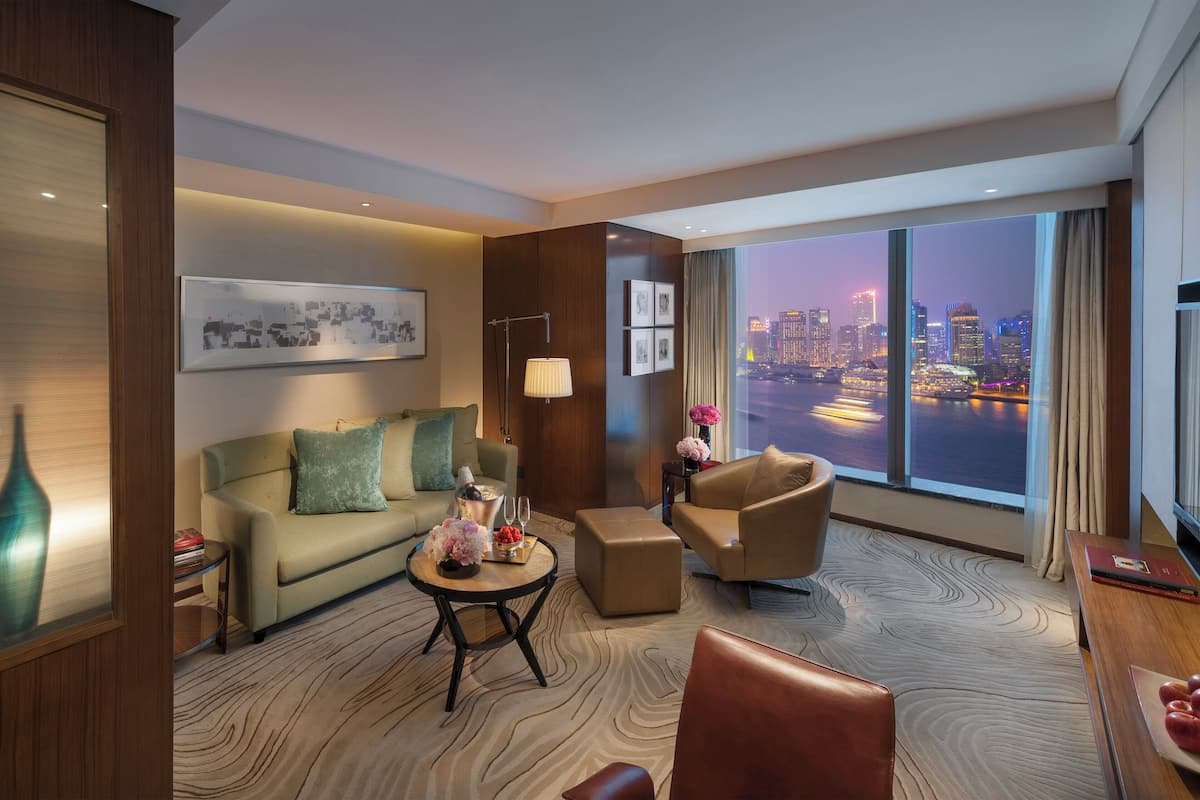 A modern hotel room with a large window offering a city view, a sofa, a coffee table, and a television.
