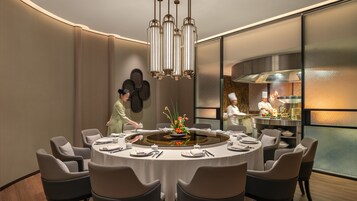 Un comedor moderno con una mesa redonda dispuesta para seis personas, un chef preparando comida al fondo y un adorno en la pared.