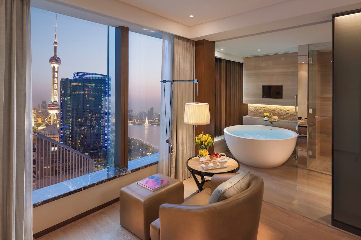 A modern hotel room with a large window offering a city view, a sitting area with a table and chairs, a bathtub, and a small kitchen area.