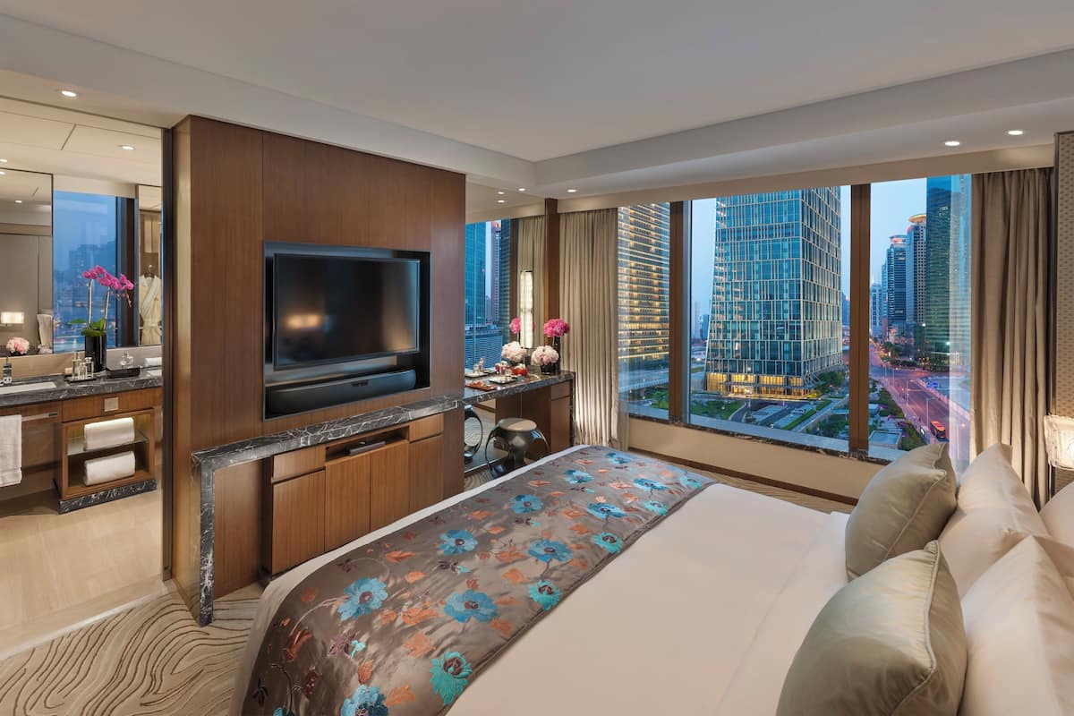 A modern hotel room with a large bed, a flat-screen TV, a desk with a chair, and a view of the cityscape through large windows.