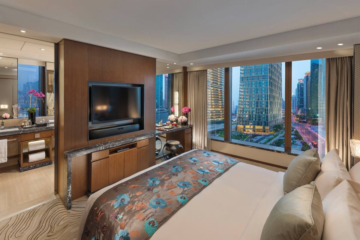 Een moderne hotelkamer met een groot bed, een flatscreen televisie, een bureau met stoel en uitzicht op de stad door grote ramen.