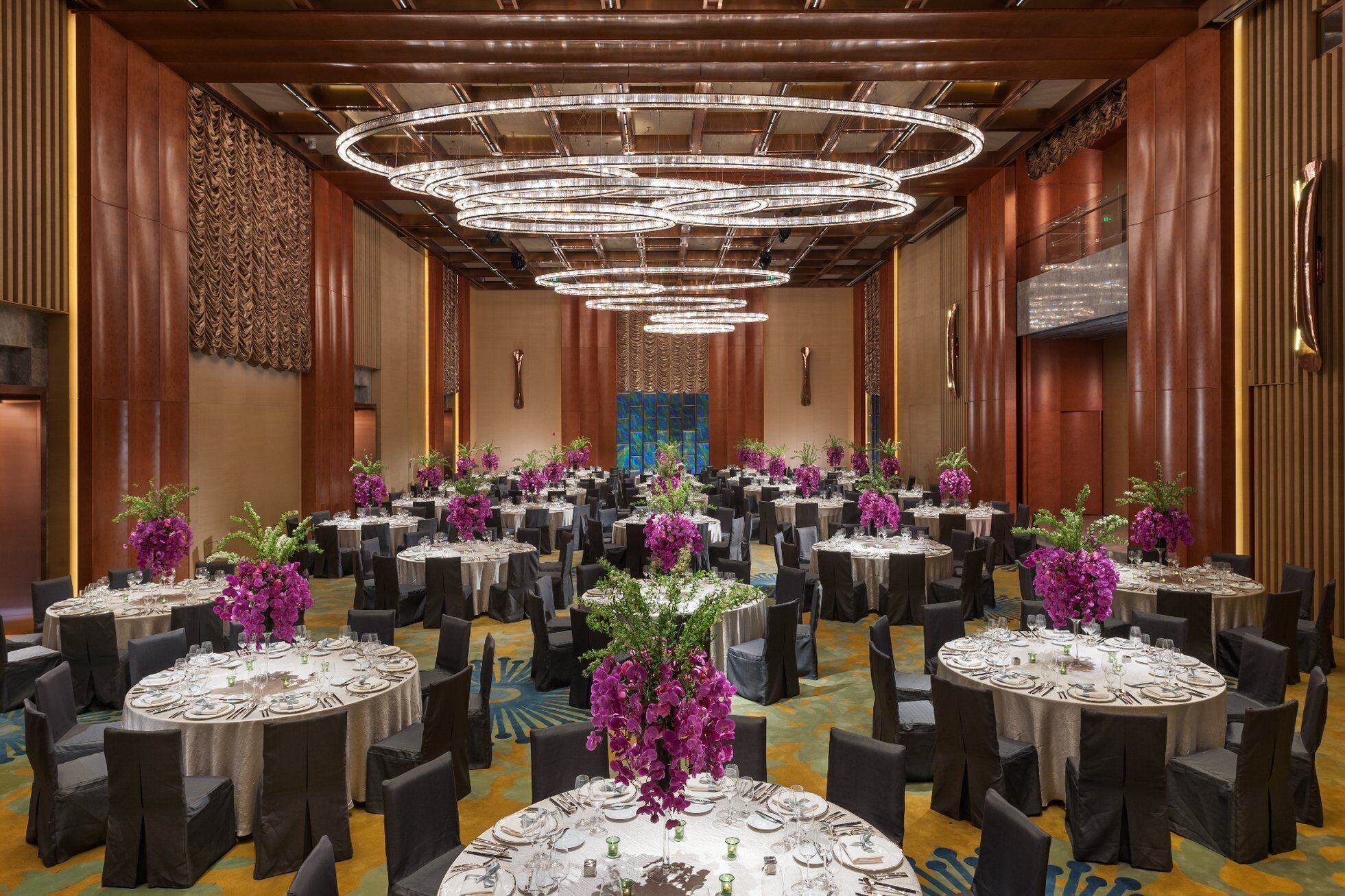 A banquet hall with multiple round tables, each set with white tablecloths, chairs, and purple flowers.