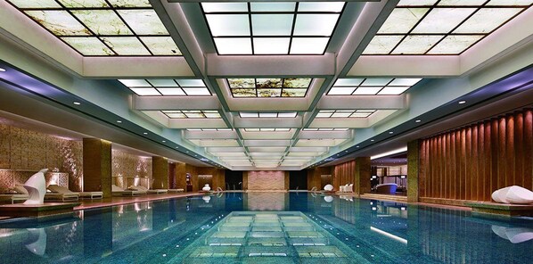 Una amplia piscina interior con techo alto y tragaluces, rodeada de zonas de descanso y una pared de piedra.