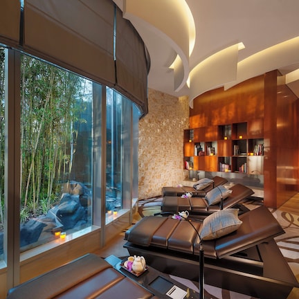 A modern spa room with a large window overlooking bamboo, a massage table, and a cozy seating area with bookshelves.
