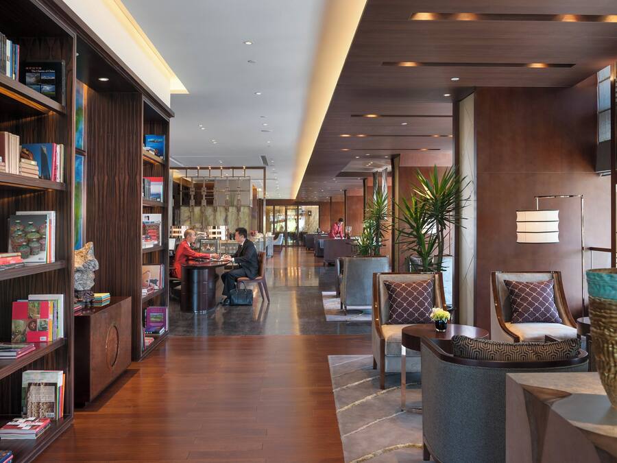 A modern hotel lobby with a long reception desk, comfortable seating areas, and a large bookshelf filled with books.