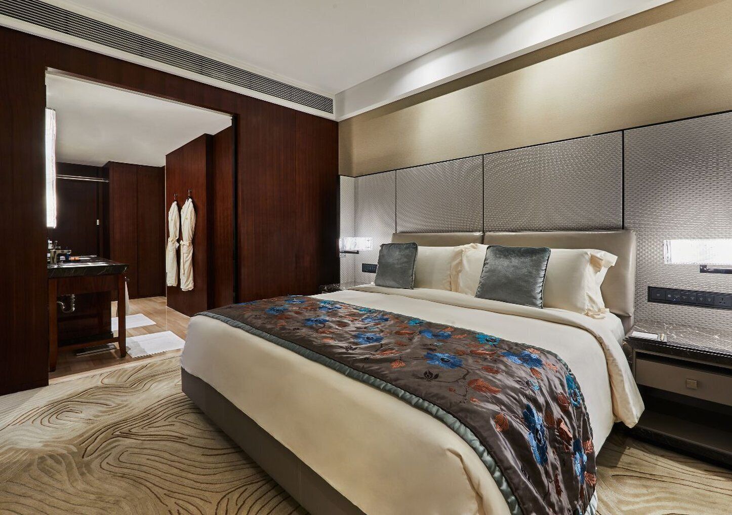 A hotel room with a large bed, a wooden wardrobe, a desk with a chair, and a bathroom visible through an open door.