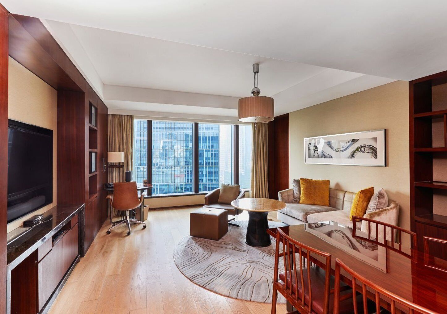 A modern hotel room with a large flat-screen TV, a dining area with a wooden table and chairs, a sofa set, and a large window offering a city view.