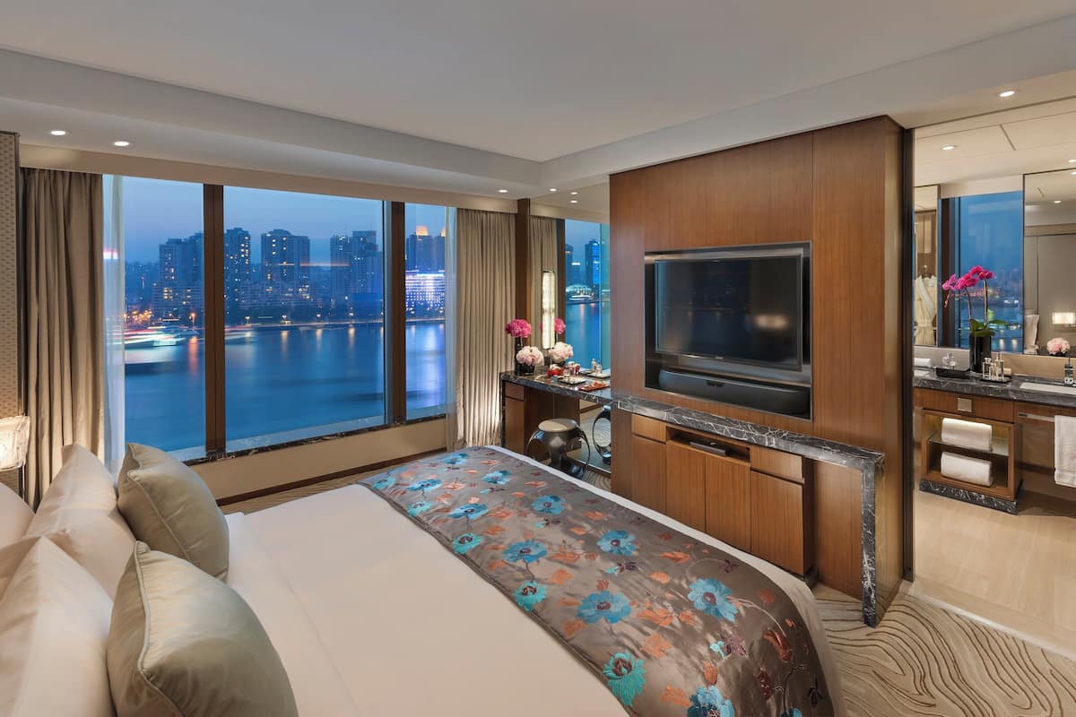 A modern hotel room with a large bed, a TV, a desk, and a view of a cityscape.