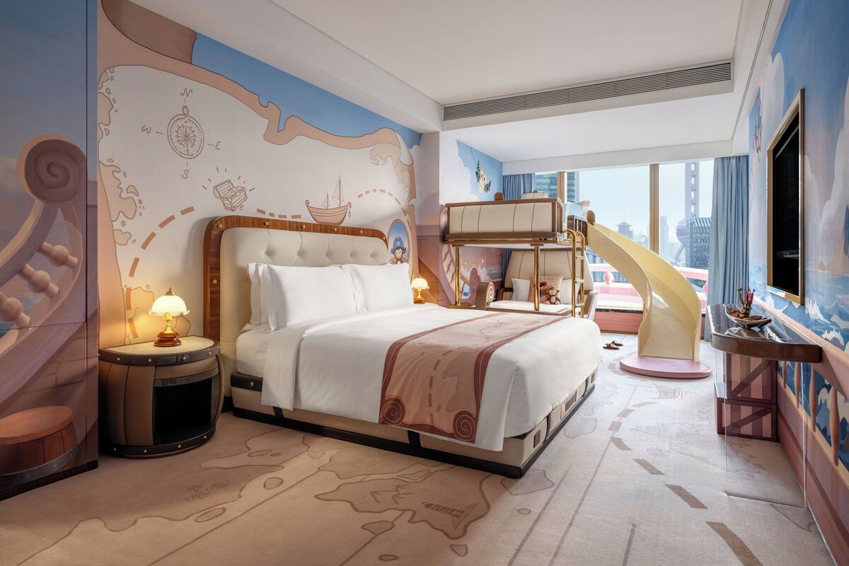 A hotel room with a large bed, a desk, a chair, a lamp, a window with a view of buildings, and a mural on the wall.
