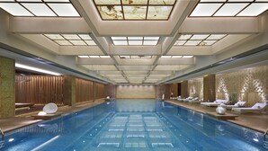 Una amplia piscina cubierta con suelo de baldosas, rodeada de zonas de descanso con tumbonas y un techo de cristal.