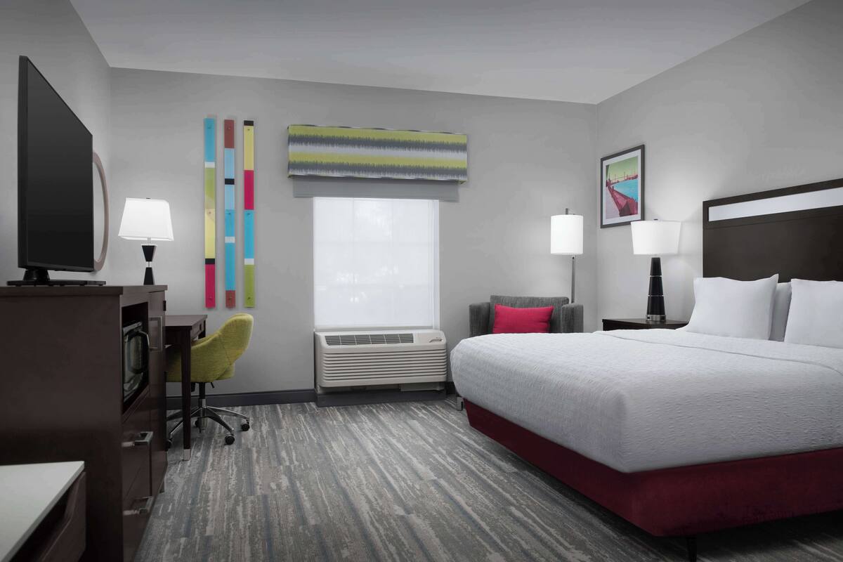 A hotel room with a large bed, a desk with a chair, a TV, a window with blinds, an air conditioning unit, and a painting on the wall.