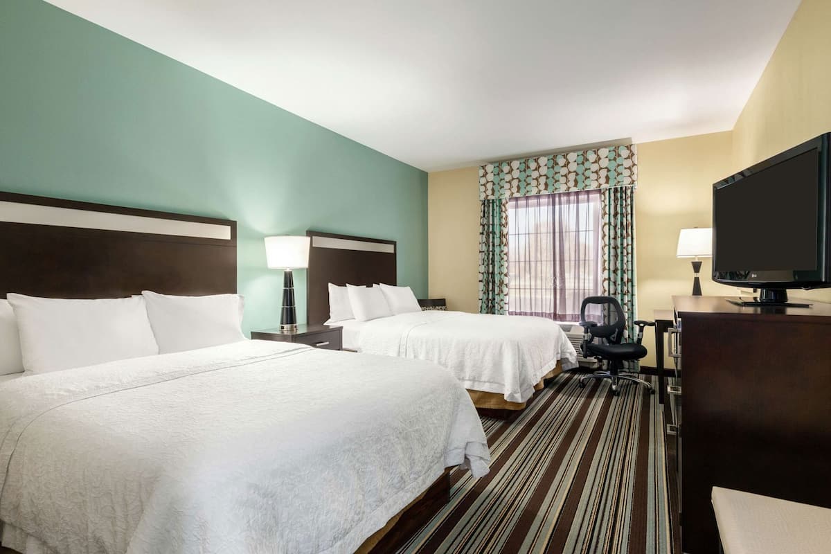 A hotel room with two beds, a desk with a television, a chair, and a window with curtains.
