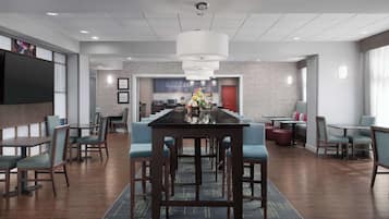 Un moderno hall de hotel con una mesa de comedor central, asientos cómodos, un televisor de pantalla plana y un área de cocina visible al fondo.