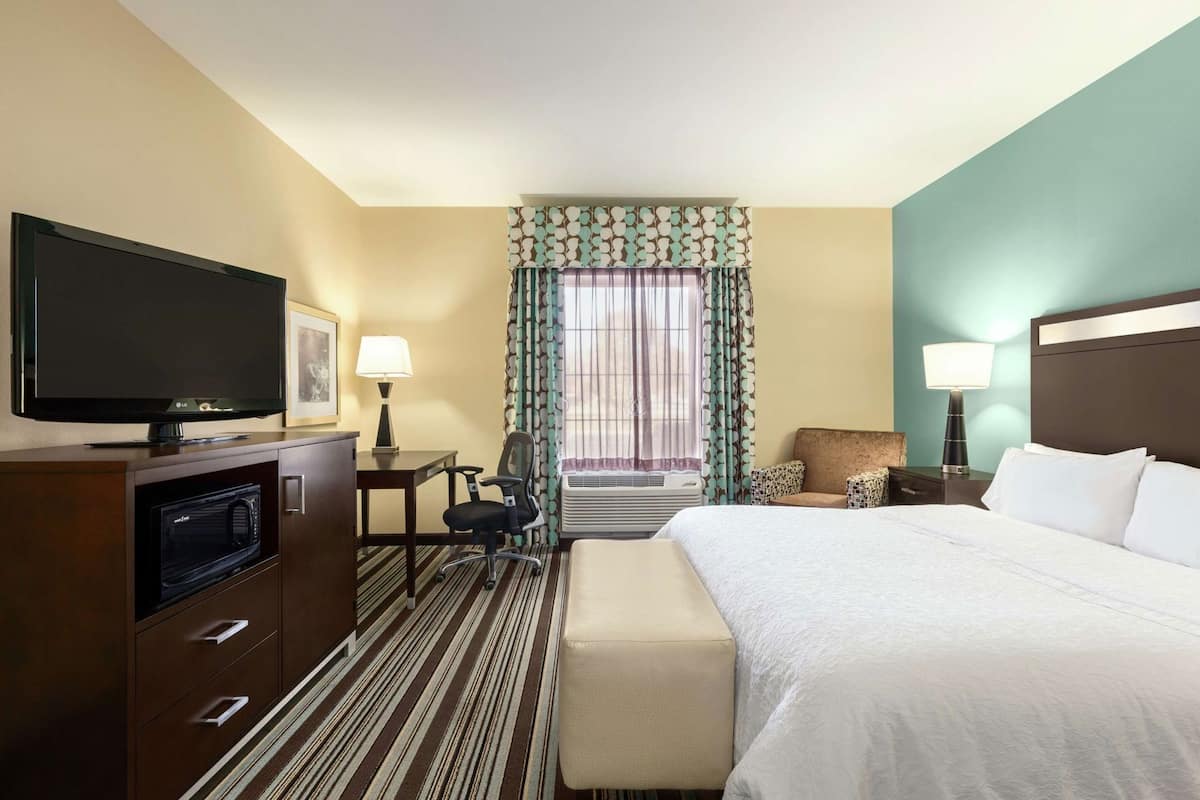 A hotel room with a bed, a desk with a lamp, a television, a microwave, and a chair.
