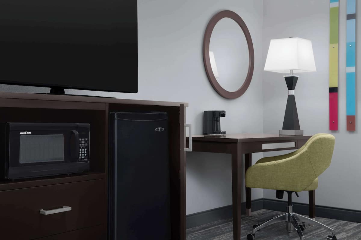 A hotel room with a flat-screen TV, a microwave, a refrigerator, a desk with a lamp, and a green office chair.