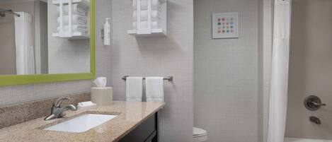 A modern bathroom with a granite countertop sink, a mirror with a green frame, two towel racks, a toilet, and a shower with a curtain.