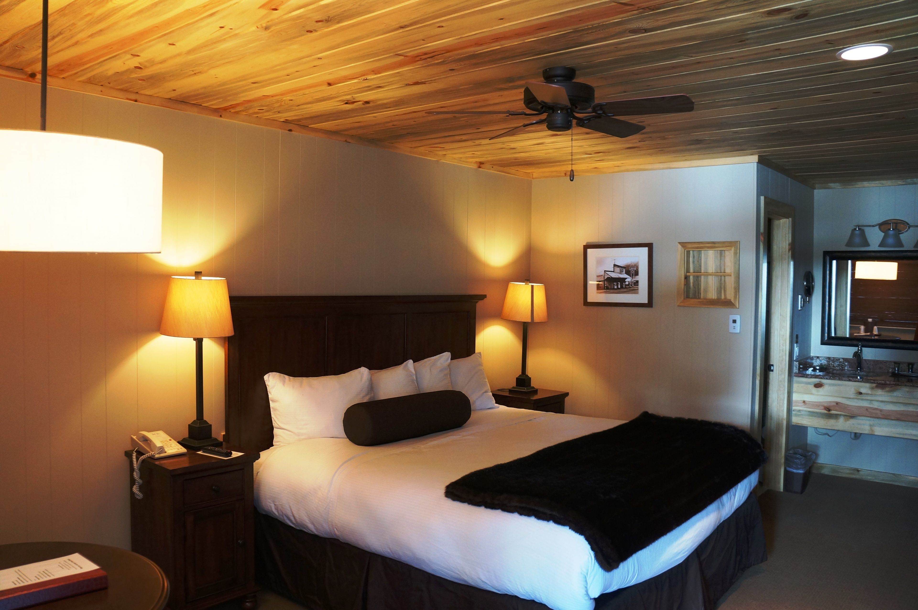 Une chambre d’hôtel avec un plafond en bois, un lit recouvert de draps blancs, deux lampes de chevet, un téléphone et une petite table sur laquelle se trouve un livre.