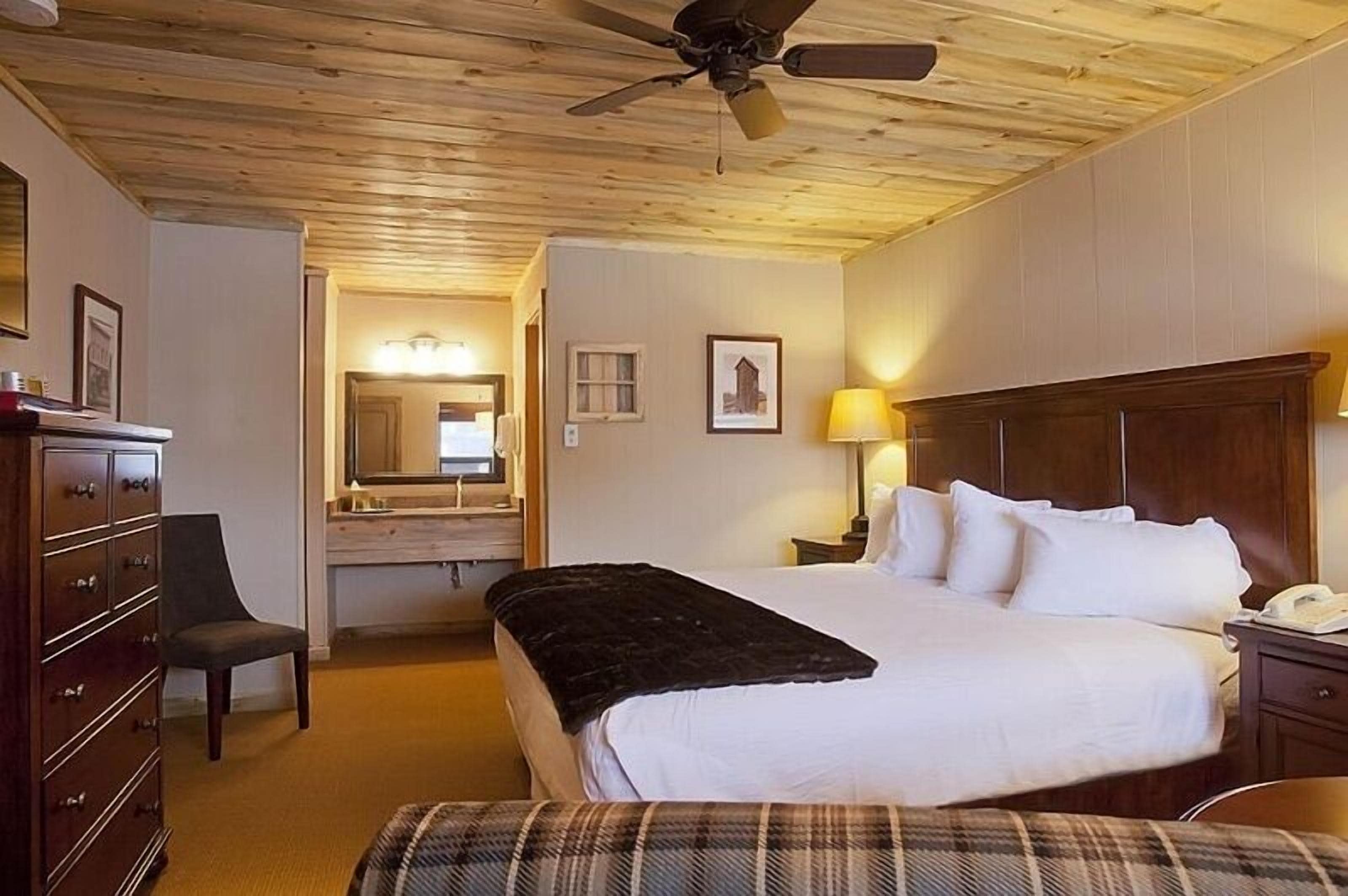 Habitación de hotel con techo de madera, cama con sábanas blancas, escritorio con lámpara, cómoda y una silla.