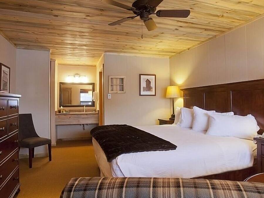 Habitación de hotel con techo de madera, cama con sábanas blancas, escritorio con lámpara, cómoda y una silla.