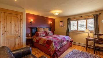 Un dormitorio con puerta de madera, una cama con colcha colorida, un sillón de cuero, una mesita de noche, una lámpara y una ventana con cortinas.