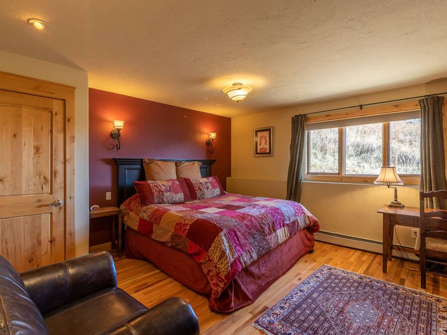 Un dormitorio con puerta de madera, una cama con colcha colorida, un sillón de cuero, una mesita de noche, una lámpara y una ventana con cortinas.