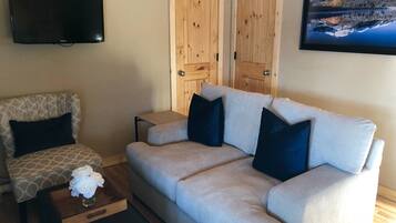 Ein Wohnzimmer mit einer Couch, einem gemusterten Sessel, einem Couchtisch mit Blumen und einem Fernseher, sowie Holztüren.