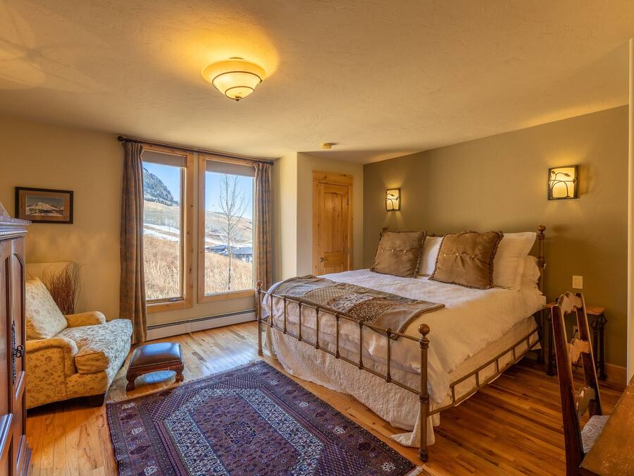 Un dormitorio con una cama grande, piso de madera, ventana con vistas al exterior, una puerta y una mesita.