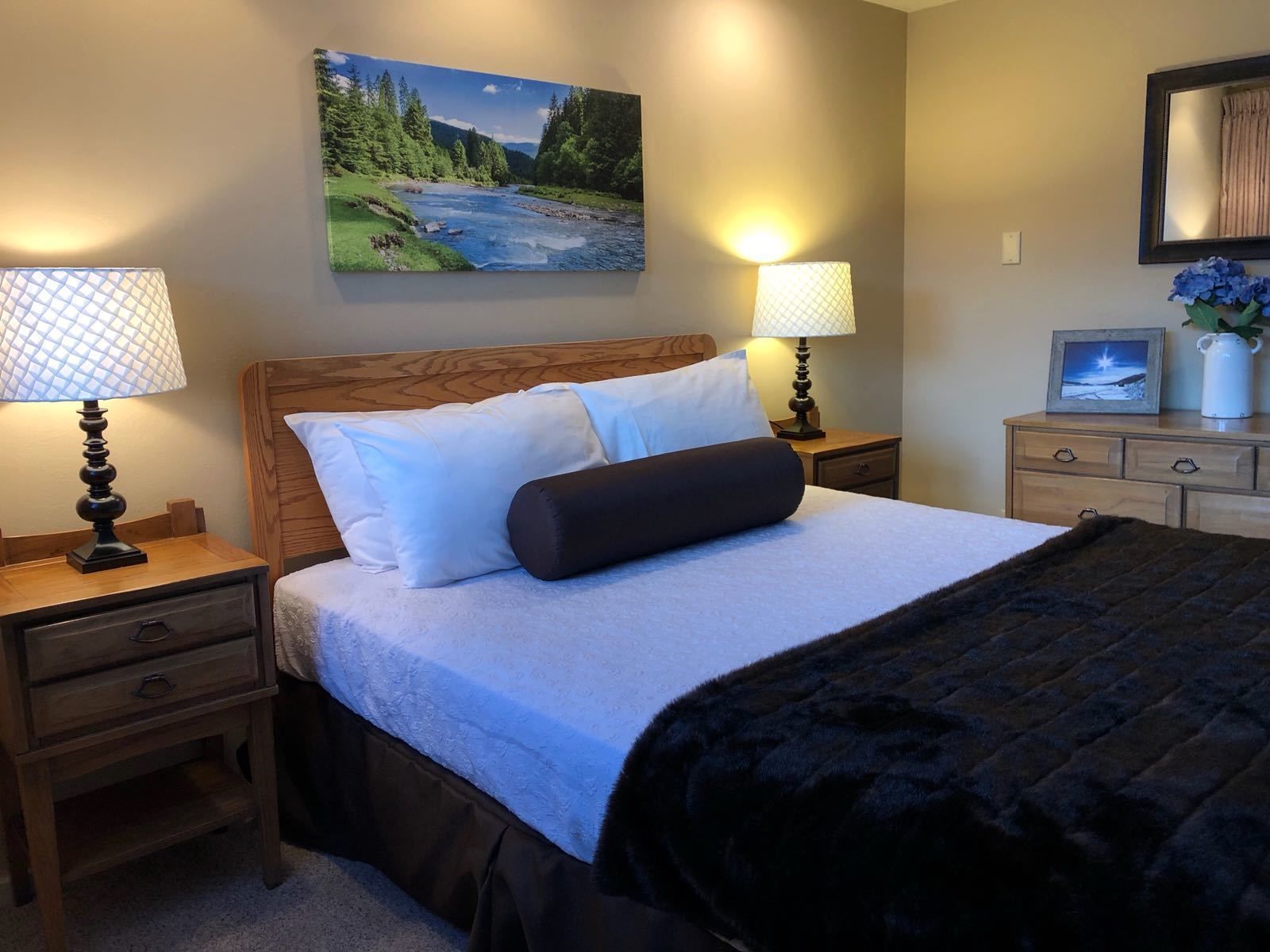 A neatly made bed with a wooden headboard, two bedside tables with lamps, a dresser with a mirror, and a painting of a river landscape on the wall.