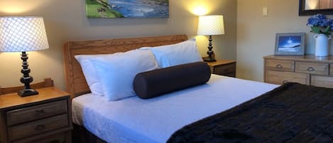 A neatly made bed with a wooden headboard, two bedside tables with lamps, a dresser with a mirror, and a painting of a river landscape on the wall.