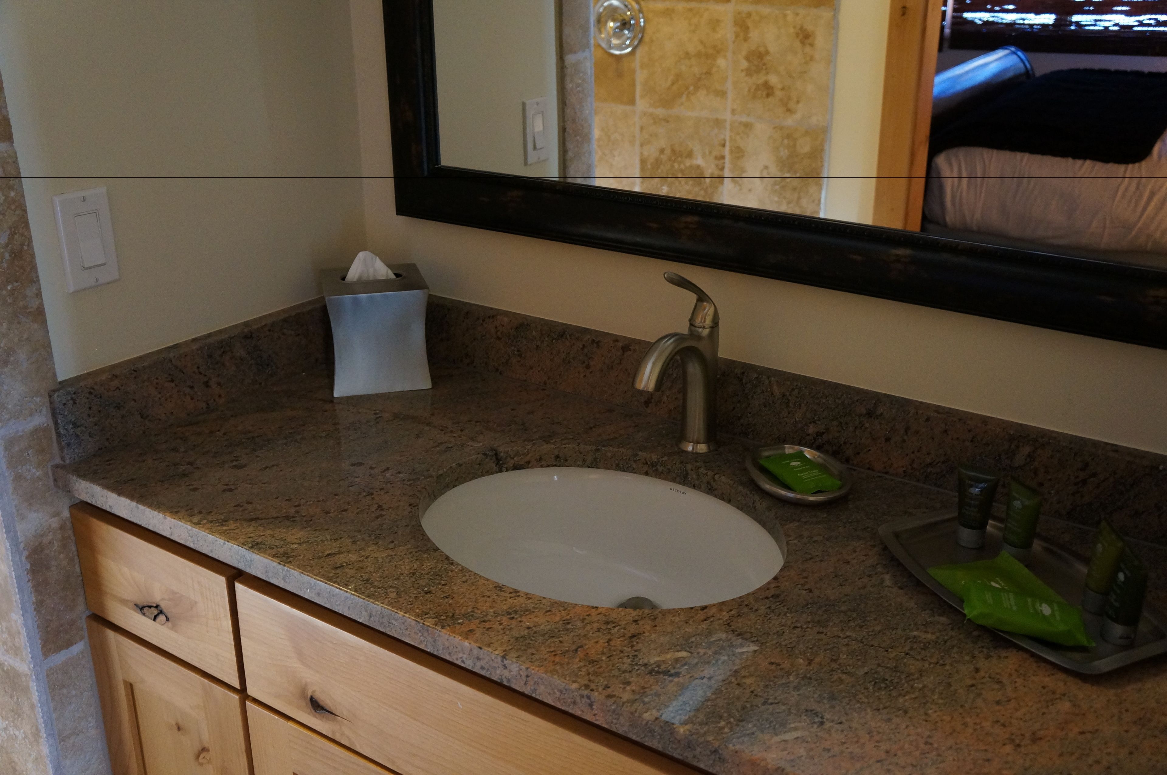 Une salle de bain avec un comptoir en granit, un lavabo blanc, un miroir, un distributeur de serviettes et des articles de toilette.