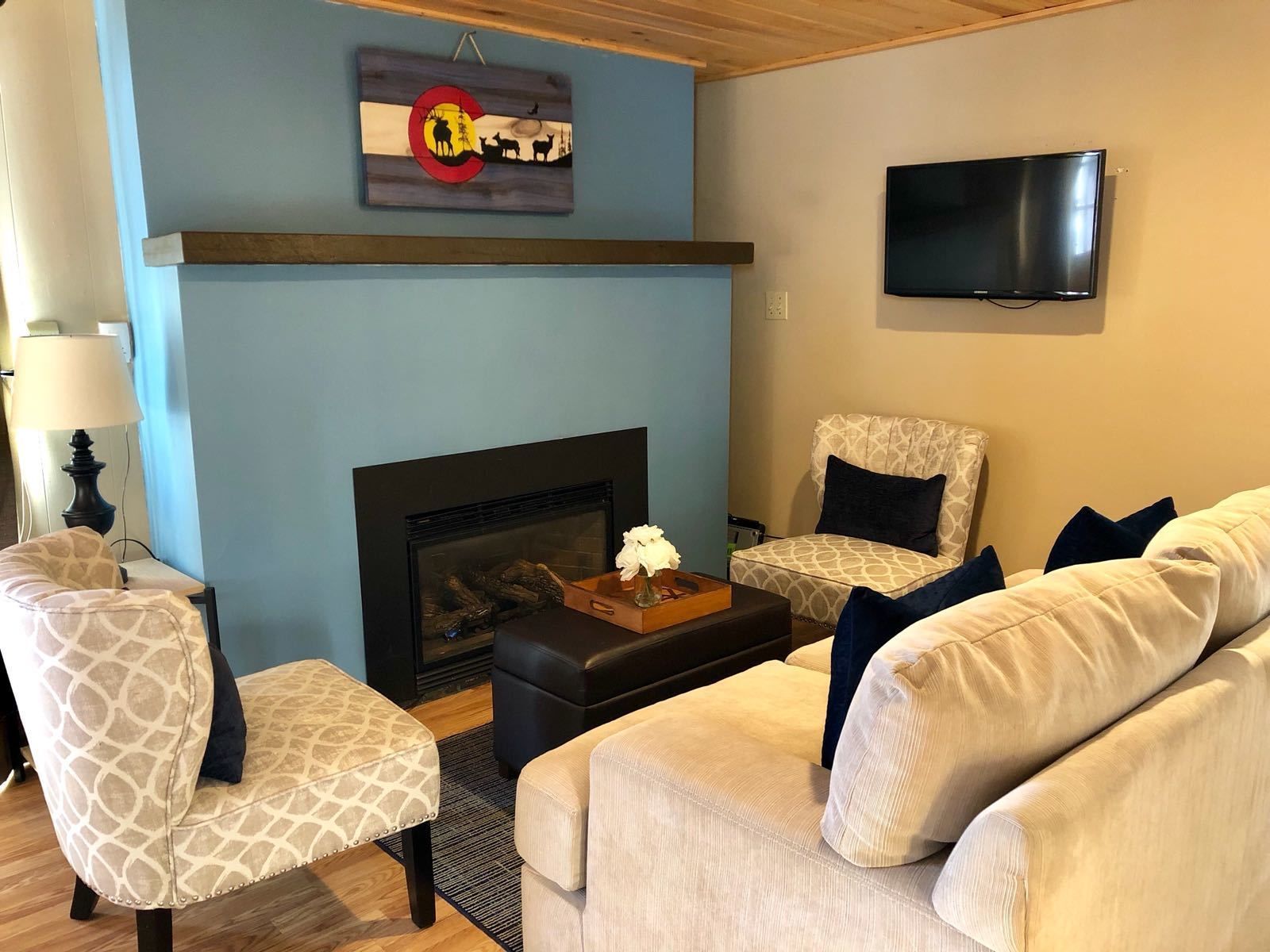 Una sala de estar con chimenea, dos sillones estampados, un sofá y una televisión en la pared.