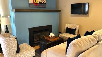 Una sala de estar con chimenea, dos sillones estampados, un sofá y una televisión en la pared.