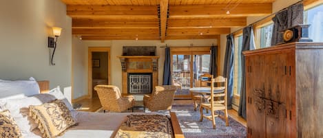 A cozy room with a wooden ceiling, a fireplace, a bed with patterned bedding, and a dining area with chairs and a table.