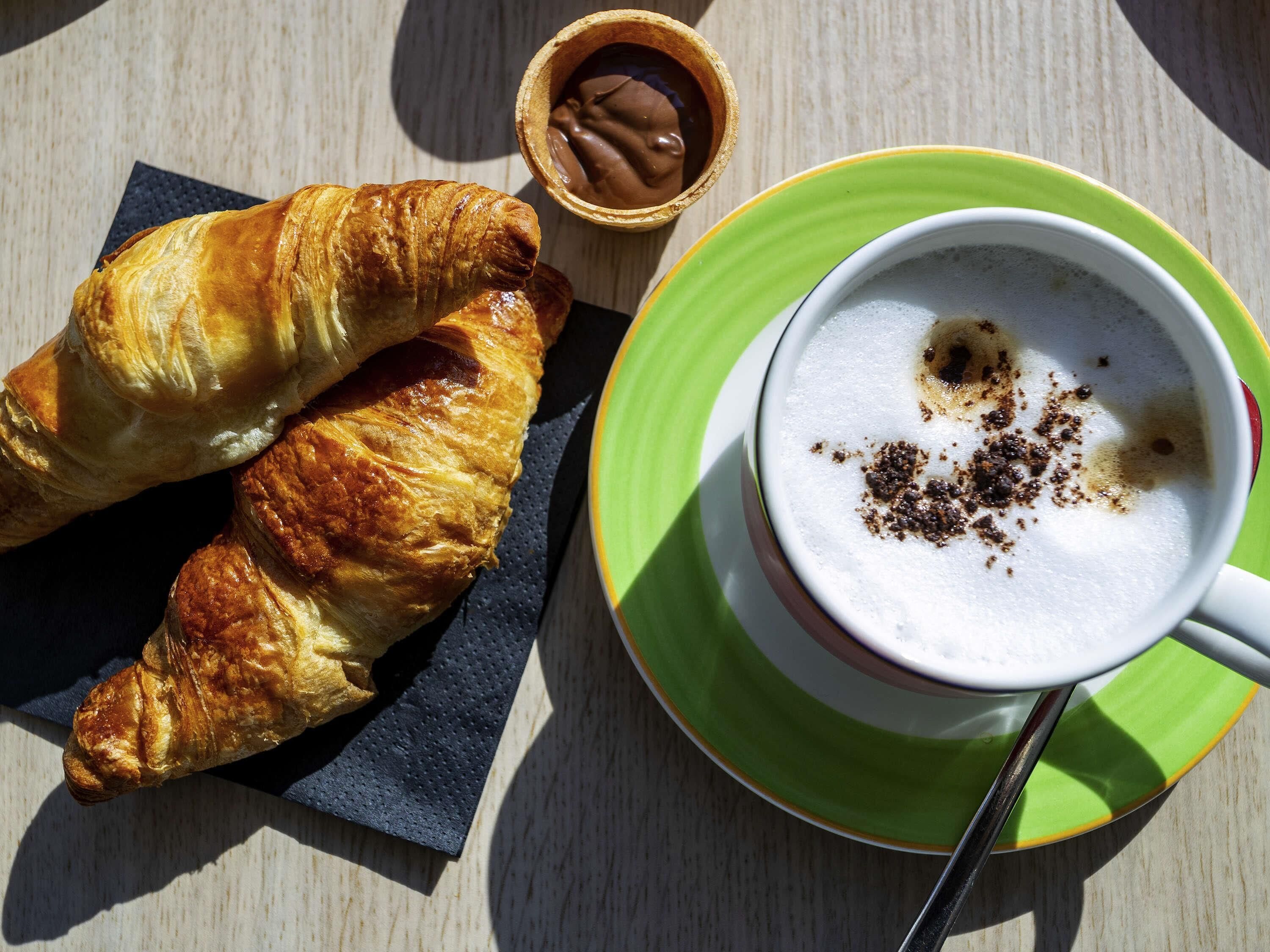 A croissant, a small chocolate container, and a cup of coffee with milk foam on a wooden table.