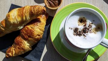 A croissant, a small chocolate container, and a cup of coffee with milk foam on a wooden table.